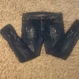 DSQUARED2 blå slitna jeans - Säljer ett par blå jeans från DSQUARED2 med coolt distressed look och slitningar framtill och baktill. Klassisk femficksmodell med normal passform och raka ben. Jeansen har en mörkblå tvätt och tydliga slitdetaljer för en edgy vibe.