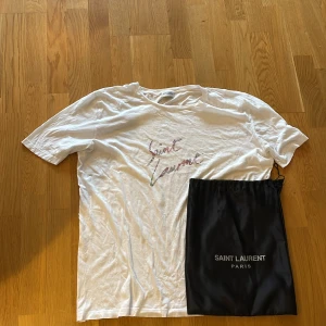 Vit Saint Laurent t-shirt - Vit t-shirt från Saint Laurent med handskriven logga i pastellfärger på bröstet. Klassisk passform med rund hals och korta ärmar. Kommer med svart dustbag med Saint Laurent Paris-tryck. Storleken är M men skulle även passa L. Hör av er vid fundering!