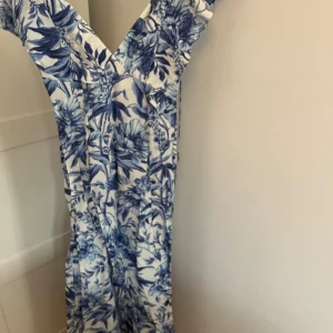 Blommig långklänning med volang - Säljer en superfin långklänning med blått och vitt blommigt mönster. Klänningen har v-ringad hals, volangdetaljer vid axlarna och knytband i midjan. Perfekt för sommarens alla tillfällen! Materialet känns lätt och luftigt.