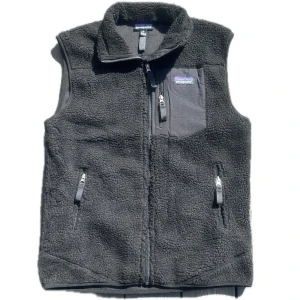 Svart fleeceväst från Patagonia - Mysig svart fleeceväst från Patagonia med hög krage och dragkedja framtill. Västen har tre fickor med dragkedja, varav en på bröstet, och är tillverkad i mjuk fleece. Perfekt för lager-på-lager och outdoor vibes.