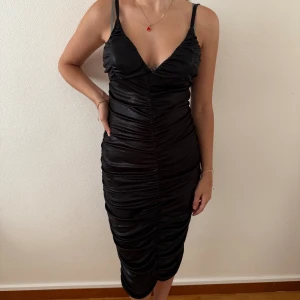 Svart omlottklänning med rynk - Snygg svart klänning med omlottkänsla och rynkade detaljer längs hela klänningen. Djup v-ringning och smala axelband ger en trendig look. Klänningen är figurnära och har en glansig finish som sticker ut.