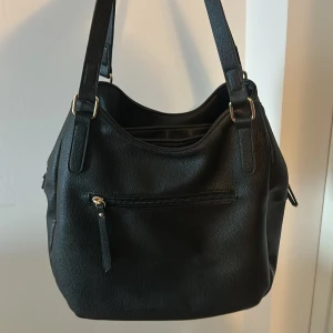 Svart handväska  - Stilren svart axelväska med guldiga detaljer och tre fack, både med dragkedja och magnetknäppning. Väskan har två breda axelband. Perfekt för dig som vill ha plats för allt du behöver i vardagen. Ordinariepris: 400