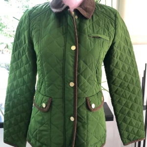 Grön quiltad jacka från Stockh LM - Quiltad grön jacka från Stockh LM med bruna manchesterdetaljer på krage och fickor. Jackan har guldiga knappar, markerad midja och lång ärm. Perfekt för dig som vill ha en klassisk men ändå unik look.