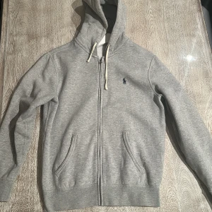 Grå hoodie från Polo Ralph Lauren - Snygg grå hoodie från Polo Ralph Lauren med dragkedja, huva med vita snören och klassisk blå logga på bröstet. Mjuk bomullskänsla och två fickor framtill. Perfekt för chill dagar eller när du vill ha en clean och enkel look. Den är använd men inga defekter eller något sådant.