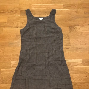 Ofice klänning  - Snygg grå klänning med rutigt mönster och två fickor framtill. Modellen är ärmlös med breda axelband och har en rak, kort passform som ger en clean och modern look. Perfekt för dig som gillar klassisk stil med en twist.