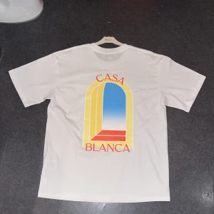 Vit Casablanca t-shirt - Vit t-shirt från Casablanca med stort färgglatt tryck på ryggen och mindre tryck på bröstet. Motivet har en gul och blå portal med röd text 'CASA BLANCA'. Klassisk rund hals och tillverkad i mjuk bomull.