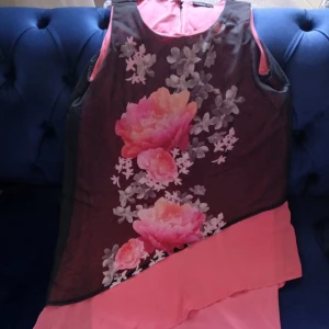 Blommig rosa kortklänning från Janina - Snygg kort klänning från Janina i rosa med svart transparent lager och stora blommor i rosa och aprikos på framsidan. Ärmlös modell med rund hals och dragkedja i ryggen. Perfekt för dig som gillar färg och blommigt. Materialet känns lätt och luftigt.