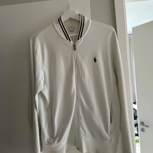 Vit kofta från Polo Ralph Lauren - Vit kofta med dragkedja från  Ralph Lauren med svart logga på bröstet och svarta ränder på kragen. Dragkedja framtill och två fickor. Skön bomullskvalitet och långärmad modell. Bra skick. Anledning till försäljning är att den är för liten för mig 