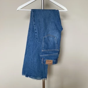 Blå jeans med rå kant - Klassiska blå jeans med rak passform och snygg rå kant nertill. Jeansen har en ikonisk bakficka och läderpatch med logga. Perfekta för en avslappnad och tidlös stil.