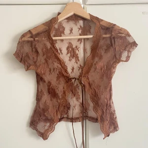 Brun spetsblus med knyt framtill - Säljer en transparent brun blus i spets med korta ärmar och v-ringning. Blusen har fina broderade blommor i tyget och knyts framtill med ett smalt band. Perfekt för lager-på-lager och ger en romantisk vibe.