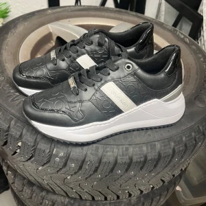 Svarta sneakers från Calvin Klein - Snygga svarta sneakers från Calvin Klein med präglad logga över hela skon. Skorna har vita och silvriga detaljer, svart snörning och en chunky vit sula. Tillverkade i skinnliknande material med modern och sportig vibe. Perfekta för dig som gillar stilrena och trendiga sneakers.