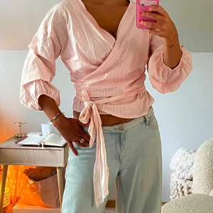 Rosa omlottblus från RUDE - Supersöt omlottblus från RUDE i ljusrosa med tunna vita ränder. Blusen har trekvartsärmar med resår och knytband i midjan som ger en snygg siluett. Perfekt för dig som gillar en feminin och trendig look.