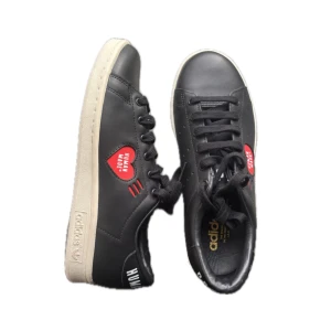 Adidas Stan Smith Human Made sneakers - Svarta Adidas Stan Smith Human Made sneakers i skinn med vit sula och röda hjärtan med texten 'Human Made' på sidorna. Klassisk rund tå, svarta skosnören och diskreta detaljer. Perfekt för dig som gillar unika samarbeten och streetstyle.