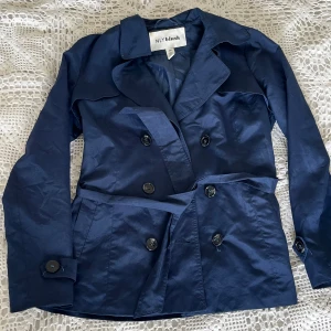 Marinblå trenchcoat  - Snygg marinblå trenchcoat i kortare modell med klassisk dubbelknäppning, bred krage och bälte i midjan. Jackan har långa ärmar och stilrena knappar framtill. Perfekt för dig som vill ha en tidlös och cool look till vardags.