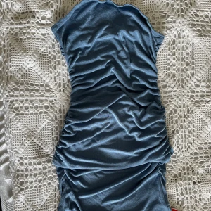 Blå tubklänning med rynkdetalj - Säljer en blå långklänning i tub-modell/offshoulder och rynkade detaljer längs hela klänningen. Modellen har en figurnära passform som framhäver siluetten. Aldrig använd med lappen kvar, OBS: scarfen som syns på andra bilden tillkommer ej då den tyvärr är borttappad. 