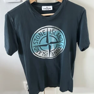 Svart t-shirt från Stone Island - Säljer en svart t-shirt från Stone Island med deras ikoniska kompasslogga i blått och silver på framsidan. Den är köpt på farfetch . På framsidan så finns det två små fläckar men som syns knappt . T-shirten är prefekt nu inför sommaren och är i bra skick ! Storlek 170 