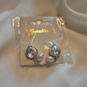 Silverfärgade örhängen med pärlor från Zanzlöza - Eleganta silverfärgade örhängen från Zanzlöza med gnistrande stenar och hängande pärlor. Perfekta för att ge en touch av glamour till din outfit. 💎✨Jag är osäker på exakt vilket material dessa är gjorda i, men de är helt oöppnade och aldrig använda. Fick dem som en present för några år sedan men glömde att jag hade dem, de är inte min stil längre men hoppas någon annan kommer gilla dem! 🥰