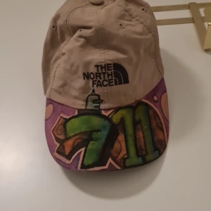 Beige keps från The North Face med graffiti - Säljer en beige keps från The North Face med unik graffiti-design i grönt och lila på skärmen. Kepsen har justerbar passform och är perfekt för en cool och avslappnad stil. Märkesloggan syns tydligt framtill.