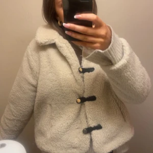 Beige teddyjacka med knappar - Säljer en mysig beige teddyjacka med svarta knappar. Jackan har en krage och långa ärmar, perfekt för våren. ❤️❤️Den är både stilren och bekväm, ett måste i garderoben!
