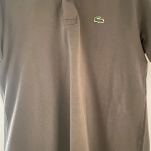 Brun pikétröja från Lacoste - Snygg brun pikétröja från Lacoste med klassisk krage och knappar framtill. Den har korta ärmar och det ikoniska gröna krokodilemblem på bröstet. Perfekt för en stilren och avslappnad look. Tveka inte på att höra av dig vid frågor eller funderingar. Mvh Sixten 