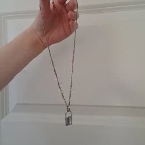 Silverfärgat halsband med hänglås - Snyggt silverfärgat halsband med ett hänglås som hänge. Kedjan är tunn och elegant, vilket gör det till en stilren accessoar. Perfekt för att ge en edgy touch till din outfit.