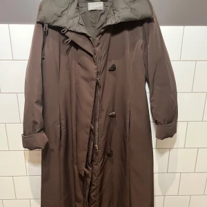 Allegri Vera Piuma D’OCA - Säljer en elegant brun kappa från Allegri. Den har en klassisk design med knappar och en hög krage. Perfekt för kyligare dagar med sin långa passform och stilrena look. Jackan är även vintage och sällsynt otroligt lätt att bära med en klassisk italiensk design som sitter snyggt