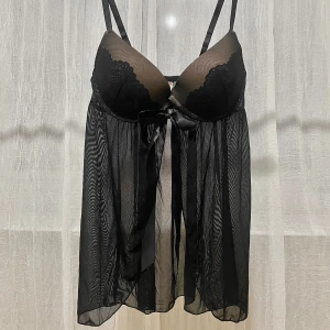 Svart negligé från Wonderland - Elegant svart babydoll från Wonderful med spetsdetaljer och en vacker rosett framtill. Den har justerbara axelband och ett transparent, flödande tyg som ger en romantisk känsla. STORLEK: 70D (EU) eller 32D (US). Passar byst 89-92cm.