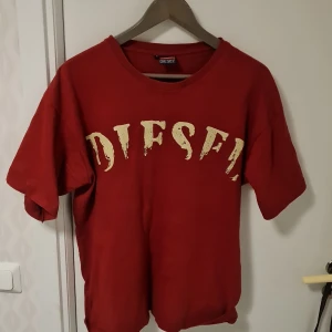 Röd t-shirt från Diesel - Säljer en röd t-shirt från Diesel med stort tryck av märkesnamnet i vitt på framsidan. T-shirten har en klassisk rund hals och korta ärmar. Perfekt för en avslappnad stil.