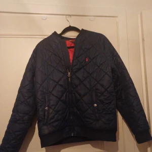 Mörkblå quiltad jacka från Ralph Lauren - Snygg mörkblå quiltad jacka från Ralph Lauren med dragkedja och två fickor med knappar. Jackan har en röd insida och en liten logga på bröstet. Perfekt för kyligare dagar!