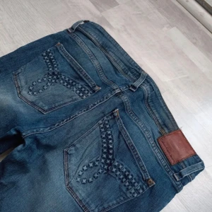 Ultra low waist blå jeans med broderade bakfickor - Ultra low waist snygga blå jeans med unika broderade detaljer på bakfickorna. De har en klassisk femficksdesign och en knappgylf. Perfekta för en avslappnad stil.