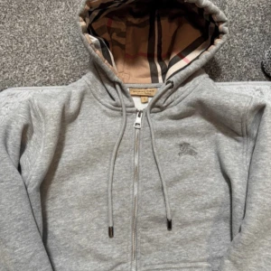 Grå hoodie från Burberry - Säljer en stilren grå hoodie från Burberry med klassiskt rutigt mönster i luvan. Tröjan har dragkedja och snörning i luvan med metalländar. Perfekt för en avslappnad och trendig look.