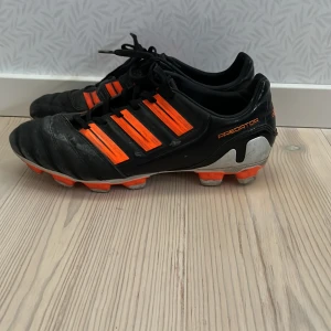 Adidas Predator fotbollsskor - Säljer ett par svarta och orangea Adidas Predator fotbollsskor med snörning. Skorna har en klassisk design med de ikoniska tre ränderna i orange på sidan och en robust sula för bra grepp på planen.