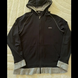 Svart kofta från Boss - Säljer en svart kofta från Boss med dragkedja och grå detaljer. Tröjan har långa ärmar och en bekväm passform. Perfekt för en avslappnad look.