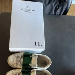 Valentino Garavani sneakers med grönt band - Snygga Valentino Garavani sneakers i vitt med ett distinkt grönt band över snörningen. Skorna har en rund tå och är tillverkade i skinn. Perfekta för en stilren look. Är även väldigt slitna så priset kan diskuteras vid snabb affär. Hör av er vid minsta fundering!
