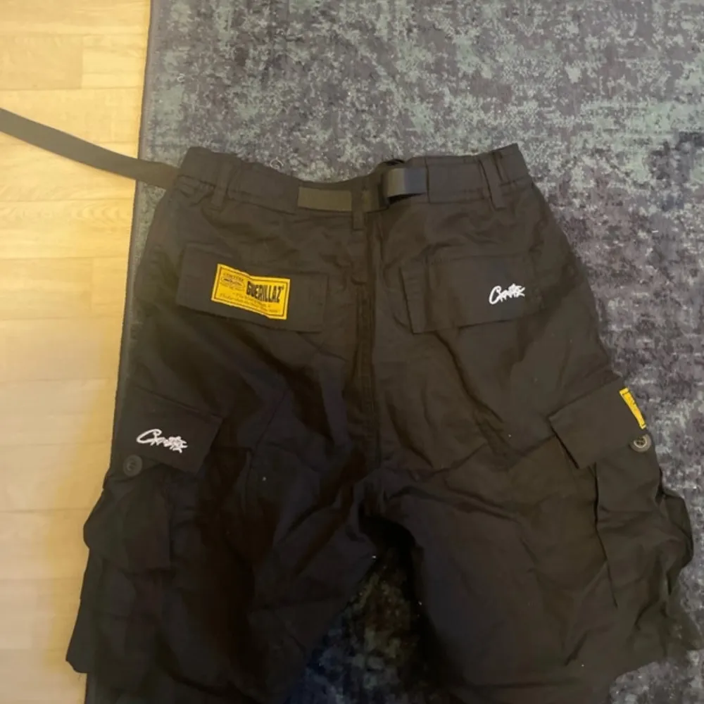 Snygga svarta shorts från Corteiz med praktiska fickor och ett coolt tryck framtill. De har en justerbar snörning i midjan och en gul etikett på sidan. Perfekta för en avslappnad stil. Aldrig använd.. Shortsit.