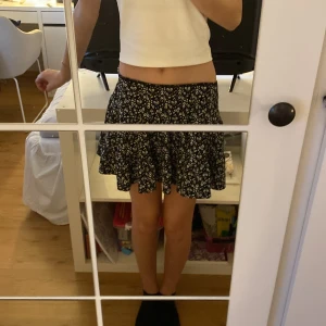 Svart blommig kortkjol - Söt svart kortkjol med ett fint blommigt mönster. Kjolen har en volangdetalj som ger den extra charm och rörelse. Perfekt för en somrig look!