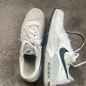 Vita Nike Air Max sneakers - Säljer ett par vita Nike Air Max sneakers med svart swoosh. Skorna har en synlig luftkudde i sulan och klassisk snörning. Perfekta för en sportig look eller vardagsstil.