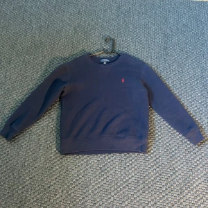 Mörkblå tröja från Polo Ralph Lauren - Säljer en stilren mörkblå tröja från Polo Ralph Lauren med det klassiska röda logotypbroderiet på bröstet.  Storlek 156-162. Tröjan har långa ärmar och en rund halsringning. Perfekt för en avslappnad och snygg look.