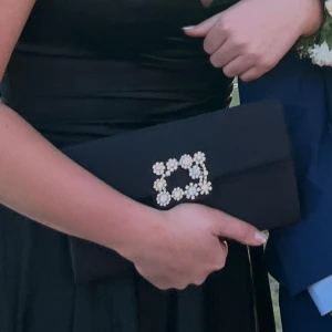 Svart kuvertväska med silver blommor  - Säljer denna superfina kuvertväskan från märket Mascara. Hade själv väskan på min studentbal och den är superfin. Finns även ett långt band till väskan som är en silverkejda. 