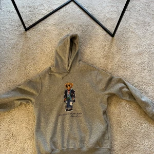 Grå hoodie från Ralph Lauren - Säljer en snygg grå hoodie från Ralph Lauren med en cool björntryck på framsidan. Hoodien har en klassisk design med huva och ribbade muddar. Perfekt för en avslappnad stil.