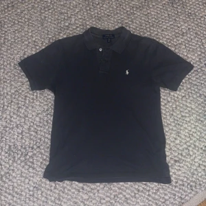 Svart pikétröja från Ralph Lauren - Snygg svart pikétröja från Ralph Lauren med klassisk krage och knappar. Den har en liten broderad logga på bröstet. Perfekt för en stilren look.