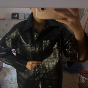 Svart skinnjacka från H&M - Snygg svart skinnjacka från H&M med krokodilpräglat mönster. Jackan har långa ärmar och knappar framtill. Stor i storleken. Hör av dig för både frågor och prisförslag!