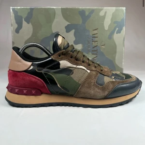 Valentino Rockrunner sneakers i camouflage - Snygga Valentino Rockrunner sneakers med ett coolt camouflagemönster. Skorna har en mix av material som mocka och skinn i färgerna grön, brun och svart. De har en unik design med röda detaljer på hälen och klassisk snörning. Endast box medföljer. Ny pris 6499