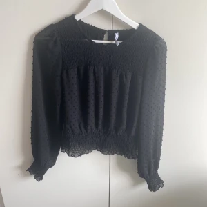 Svart blus från new Yorker  - Snygg svart blus från FB Sister med prickigt mönster och smockdetaljer. Blusen har långa ärmar och en elastisk midja som ger en fin passform. Perfekt för en stilren look.