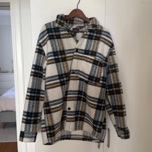 Rutig overshirt från H&M - Säljer en stilren rutig overshirt från H&M i beige, blå och gul. Overshirten har långa ärmar och knappar framtill. Perfekt för lager-på-lager under kyligare dagar.