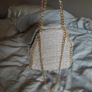 Ljust beigegrå virkad väska med guldkedja - Snygg beigegrå virkad väska med en elegant guldkedja. Väskan har en unik textur och är perfekt för att ge din outfit en trendig touch. Perfekt för att bära dina nödvändigheter med stil.