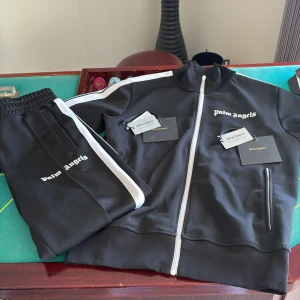 Svart tracksuit från Palm Angels - Snygg svart tracksuit från Palm Angels med vita detaljer och dragkedja framtill. Jackan har en hög krage och vita ränder längs ärmarna. Perfekt för en sportig look eller avslappnad stil. Äkta såklart och kan även säljas separat 🤗🤗