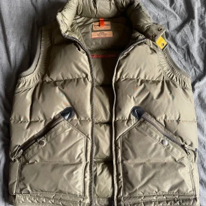 Olivgrön dunväst från Parajumpers - Säljer en olivgrön dunväst från Parajumpers Masterpiece Series. Västen har en hög krage och dragkedja framtill. Två stora fickor med dragkedja. Perfekt för kyliga dagar. Säljer denna då den inte kommit till använding använd ett fåtal gånger