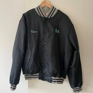 Svart bomberjacka från Rennoc, L - Retro svart bomberjacka från Rennoc med broderad text 'Vince' och siffran 24 på bröstet. Jackan har grå- och svart-randiga muddar, knäppning med svarta knappar och två fickor. Insidan är quiltad i vitt för extra komfort. Perfekt för dig som gillar sportig vintage vibe.