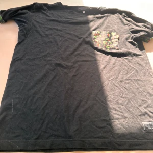 Svart t-shirt med blommig ficka SQRTN - Svart t-shirt från SQRTN Company i storlek S. Har en blommig bröstficka och matchande blommigt tyg på insidan av ärmsluten. Finns tecken på användning är lite nopprig men kan nog försvinna med noppborttagare.